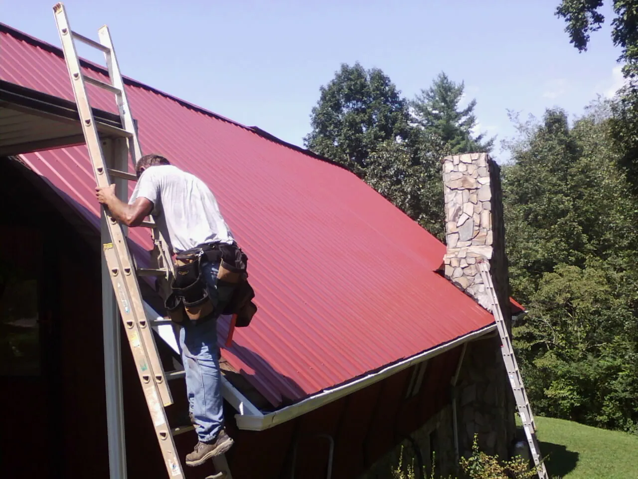 Roofer on a ladder beside a red metal roof and stone chimney — Soffit & Fascia Repair by Roofing Brothers in Tingley